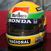 Kleurenfoto van Senna's helm, overwegend geel, met een groene streep en sponsors, in het Honda Museum.