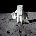 Fotografie colorată a lui Aldrin într-un costum lunar care transportă cutii cu experimente care vor fi efectuate pe lună.