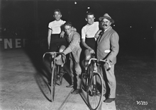 Fotografie alb-negru care prezintă doi bicicliști pe biciclete, staționari pe o pistă, sprijinindu-se pe doi bărbați îmbrăcați în civil.